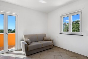 A room with a grey sofa, a sliding glass door, and a window with a view of greenery.