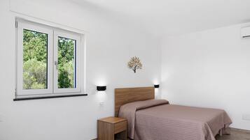 Een slaapkamer met een bed, nachtkastje, wandlamp en een raam met uitzicht op bomen.