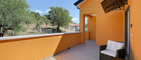 A balcony with a wicker sofa and a table, overlooking a fenced area with trees and a building in the background.