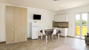 A compact kitchen with a dining area, a flat-screen TV mounted above the counter, and a sliding door leading to a balcony.