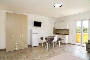 A compact kitchen with a dining area, a flat-screen TV mounted above the counter, and a sliding door leading to a balcony.