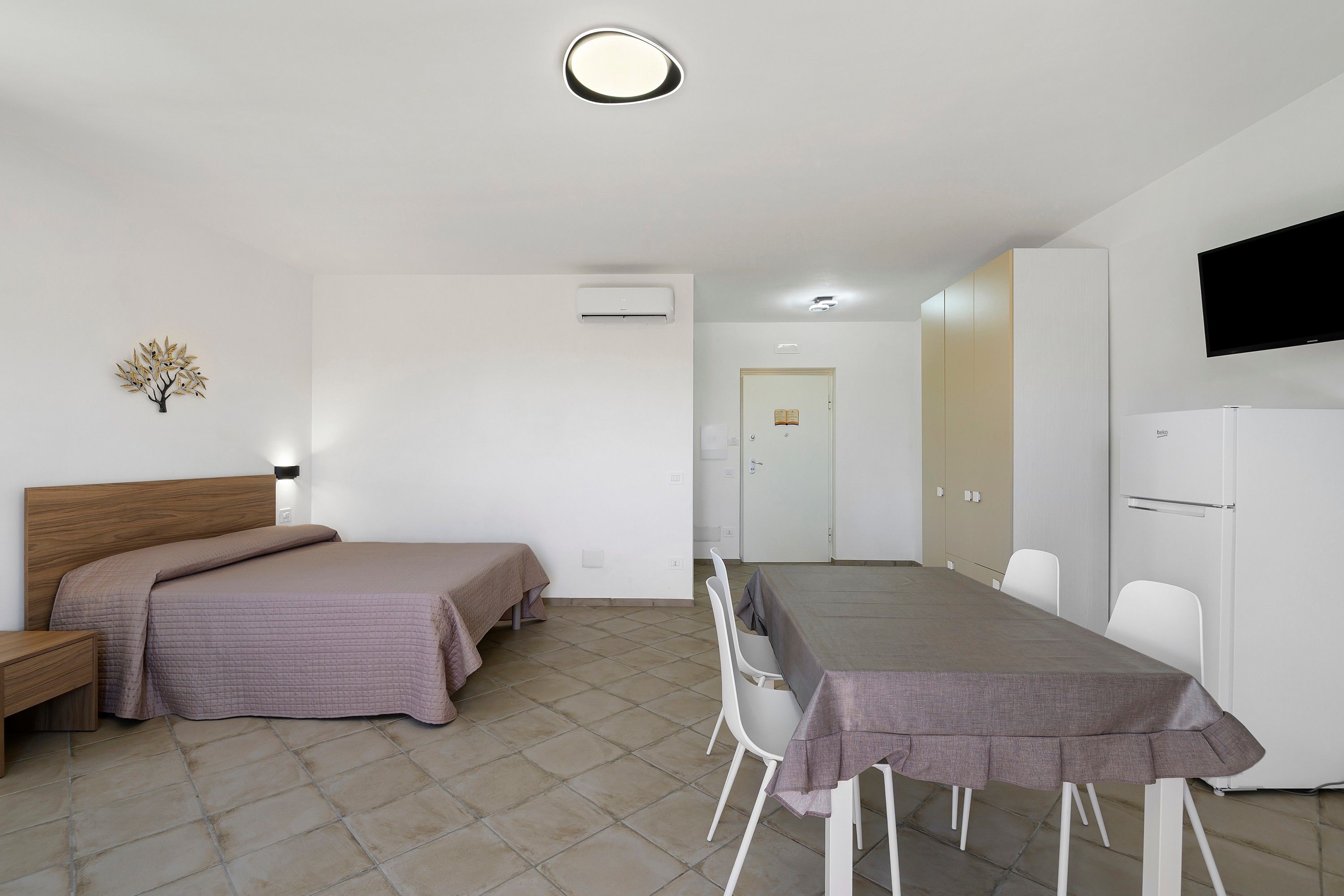 Una camera d'albergo con un letto, un comodino, una scrivania con sedie, una televisione e un frigorifero.