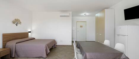 A hotel room with a bed, bedside table, a desk with chairs, a television, and a refrigerator.