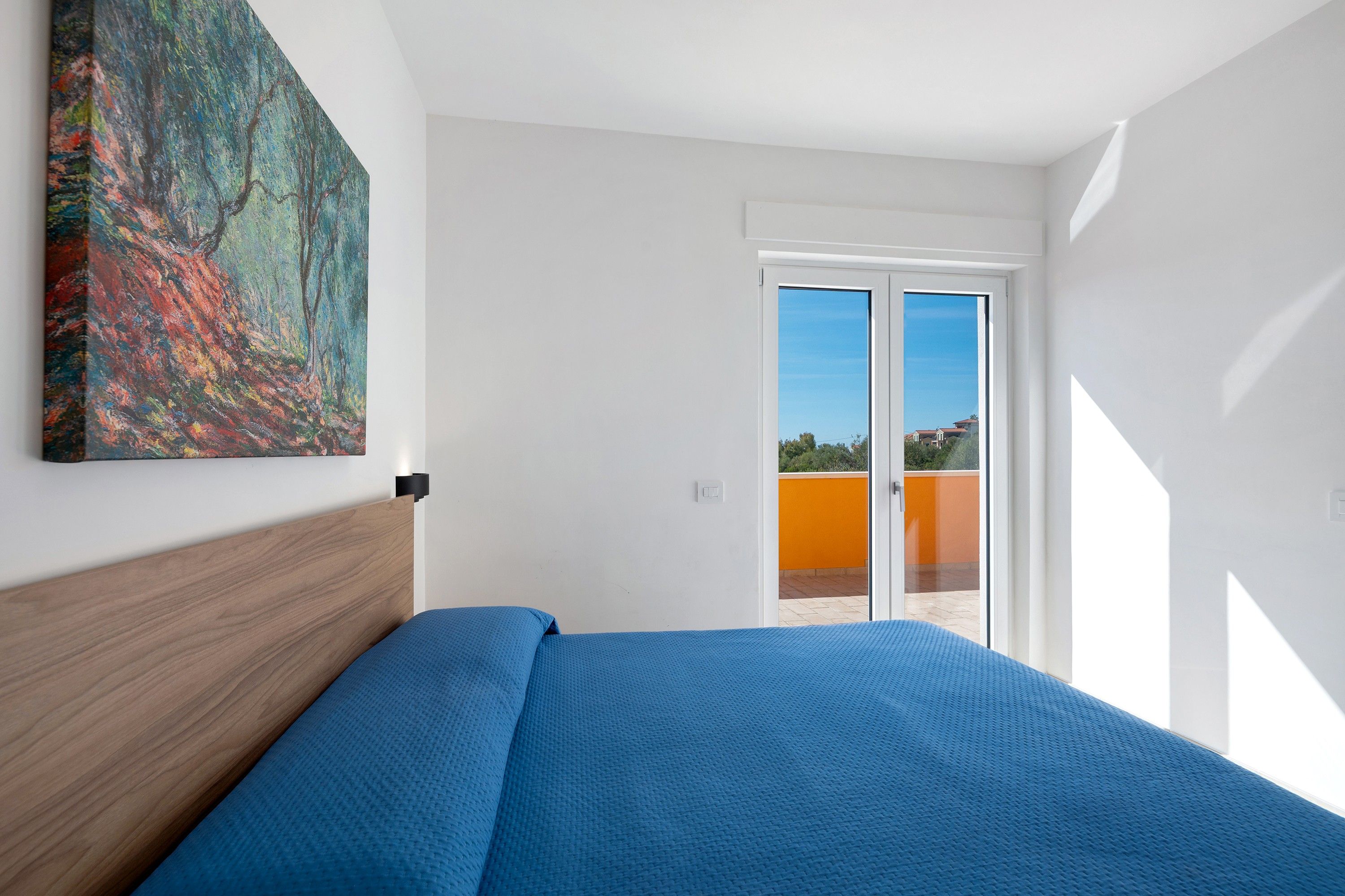 A bedroom with a bed, a painting, and a view through a door to an outdoor area.