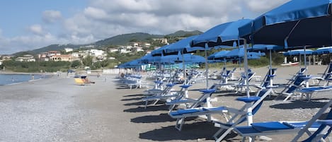 A beach with rows of blue sun loungers and umbrellas, a rocky shoreline, and buildings in the background.