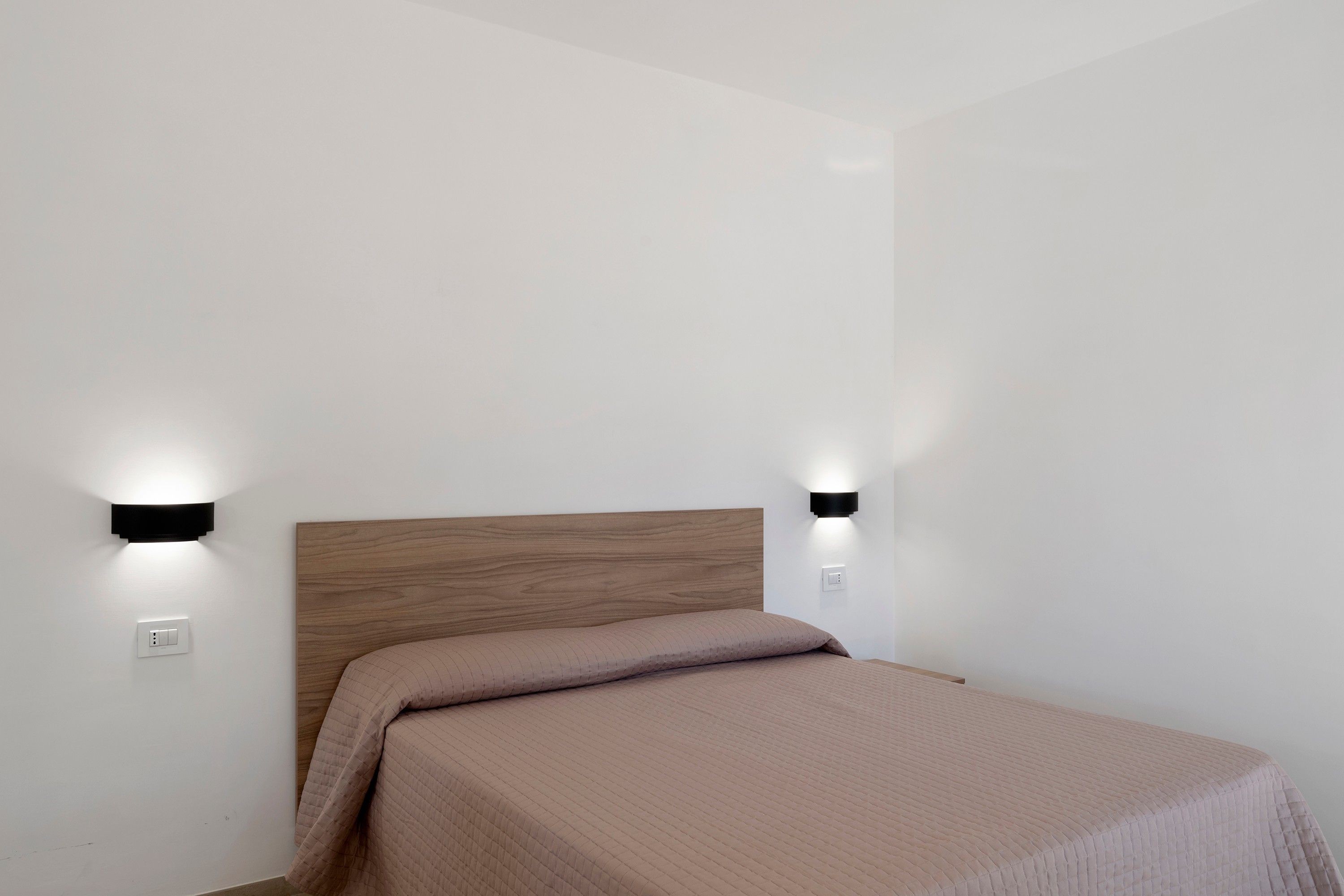 Un letto singolo con una testiera in legno in una stanza dalle pareti bianche e due luci a parete.