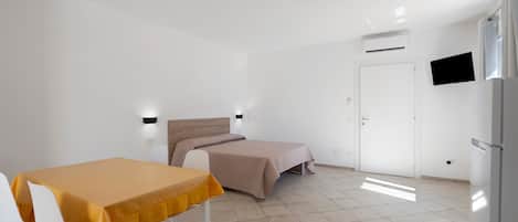 Una habitación pequeña y moderna con una cama, una mesa con sillas, un televisor en la pared y una puerta.