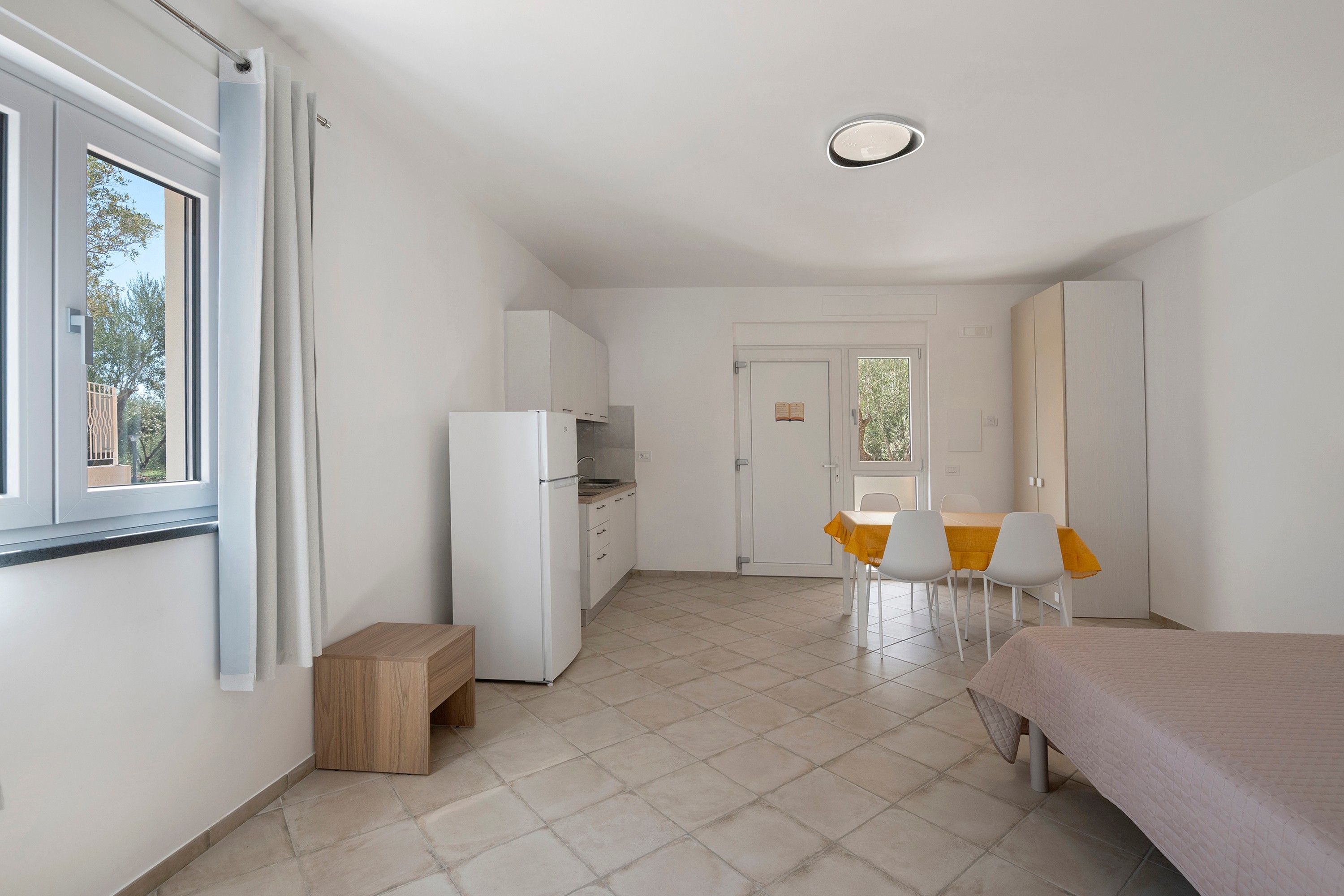 Une pièce de taille modeste comprenant un lit, une petite kitchenette, un coin repas avec une table et des chaises, et une porte donnant sur une autre pièce.