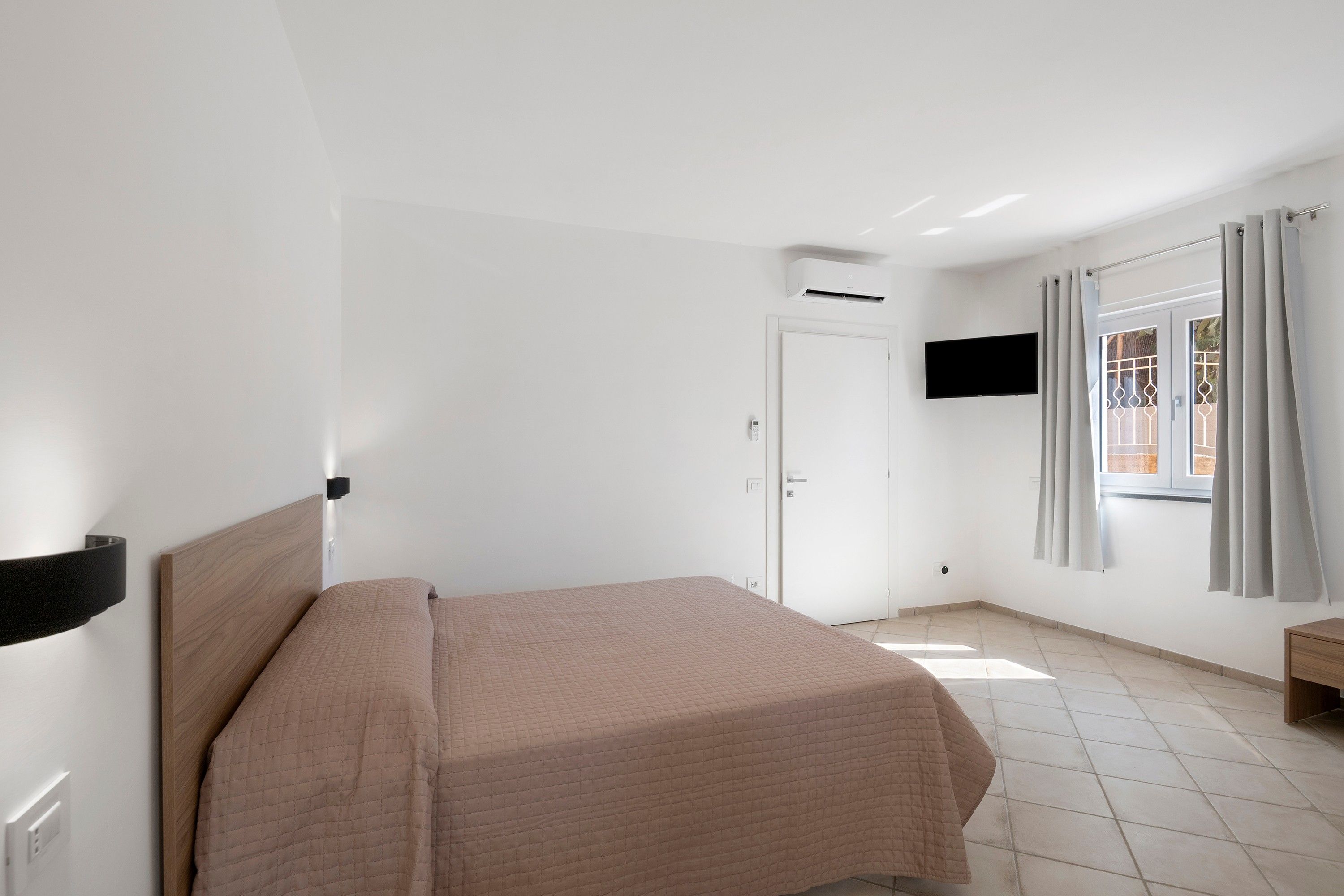 Une chambre d’hôtel avec un lit, un téléviseur fixé au mur, un appareil de climatisation, une porte et une fenêtre avec des rideaux.