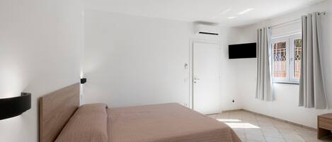 A hotel room with a bed, a TV mounted on the wall, an air conditioning unit, a door, and a window with curtains.