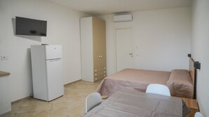 Ein kleiner, schlicht eingerichteter Raum mit einem Bett, einem Schreibtisch, einem Stuhl, einem Kühlschrank, einem Kleiderschrank und einem an der Wand befestigten Fernseher.