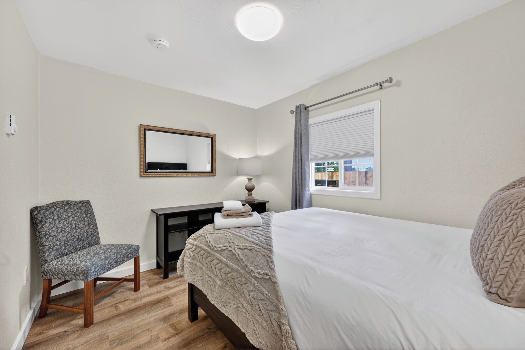 Une chambre à coucher comprenant un lit, une chaise, un bureau, une lampe, une fenêtre avec des rideaux et un miroir sur le mur.