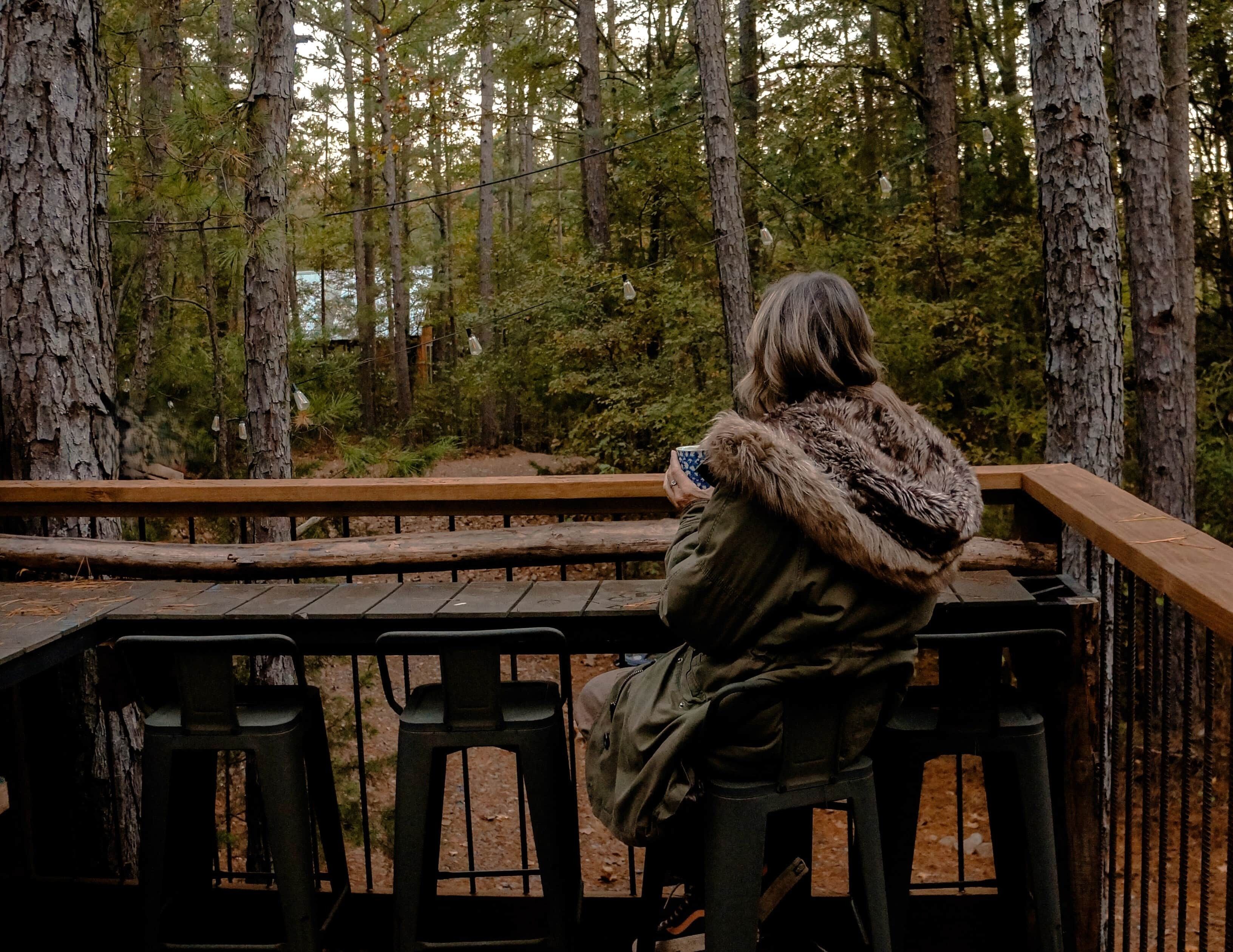 Une personne est assise sur un banc, d’où elle a une vue sur une zone boisée, avec une terrasse en bois et des garde-corps.