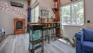 Uma sala de estar aconchegante com lareira, um espaço de bar com banquetas, uma janela grande com vista para árvores e um sofá azul.