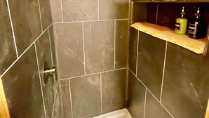 Uma casa de banho com paredes revestidas a azulejos escuros, zona de duche e uma prateleira de madeira com artigos de higiene pessoal.