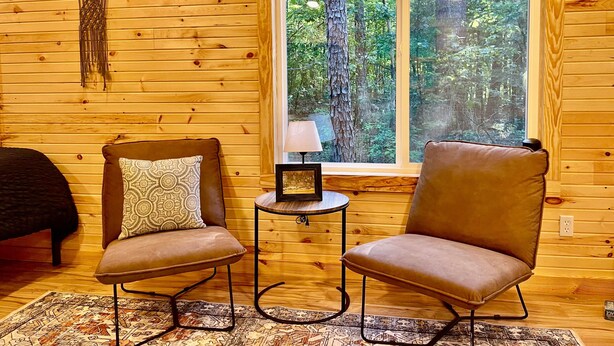 Interior acolhedor de uma cabana com duas poltronas de couro, um tapete estampado e uma janela com vista para uma floresta.