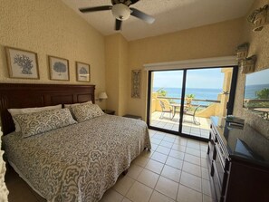A hotel room with a bed, a ceiling fan, a large window with a view of the ocean, and a TV on the right side. - Peaceful 2BR/2BA condo with Wrap-Around patio that overlooks the Sea of Cortez. (Cabo San Lucas)