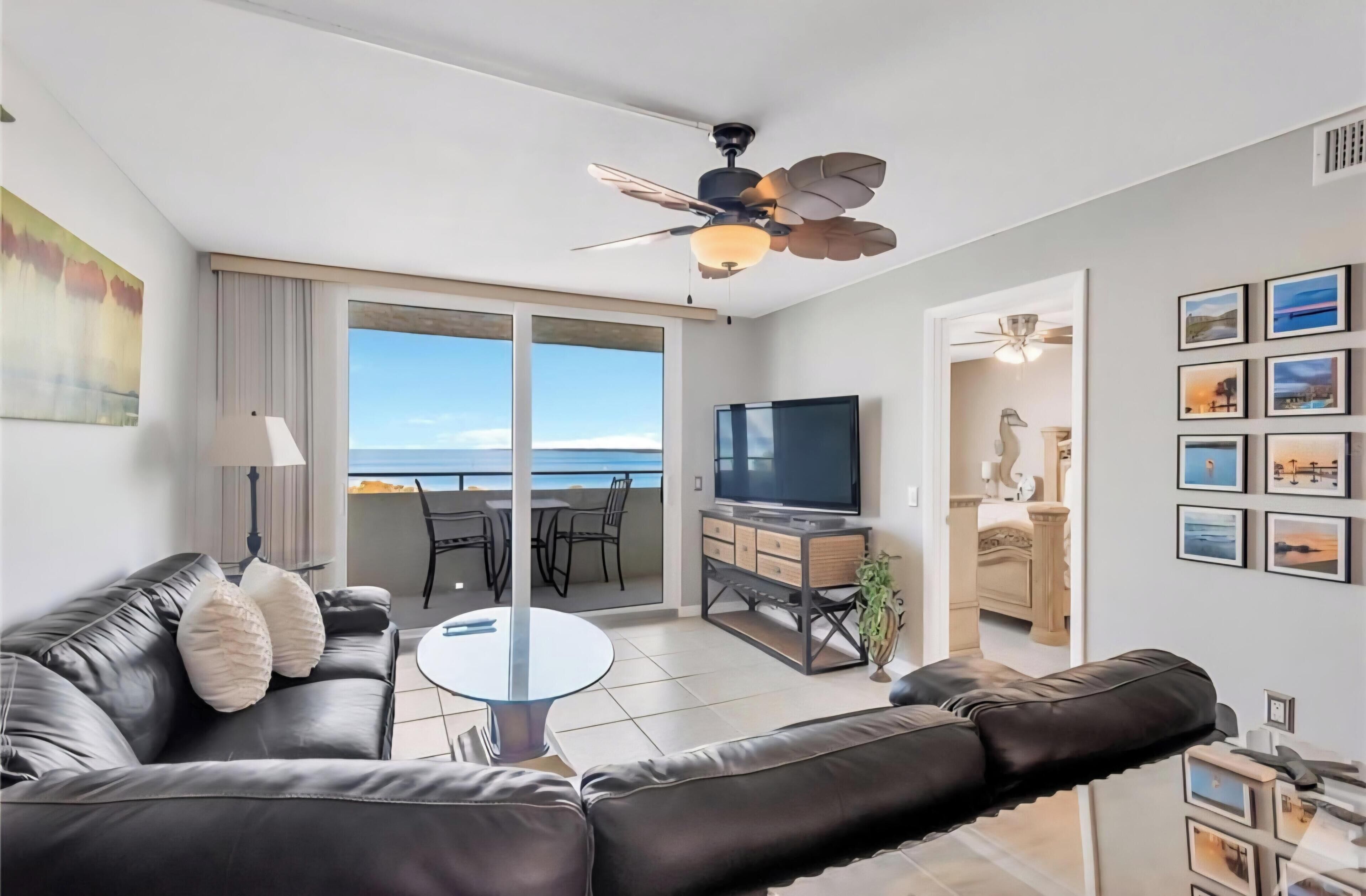 Een moderne woonkamer met een leren bank, een flatscreen televisie, een eethoek met stoelen en een balkon met uitzicht op zee.