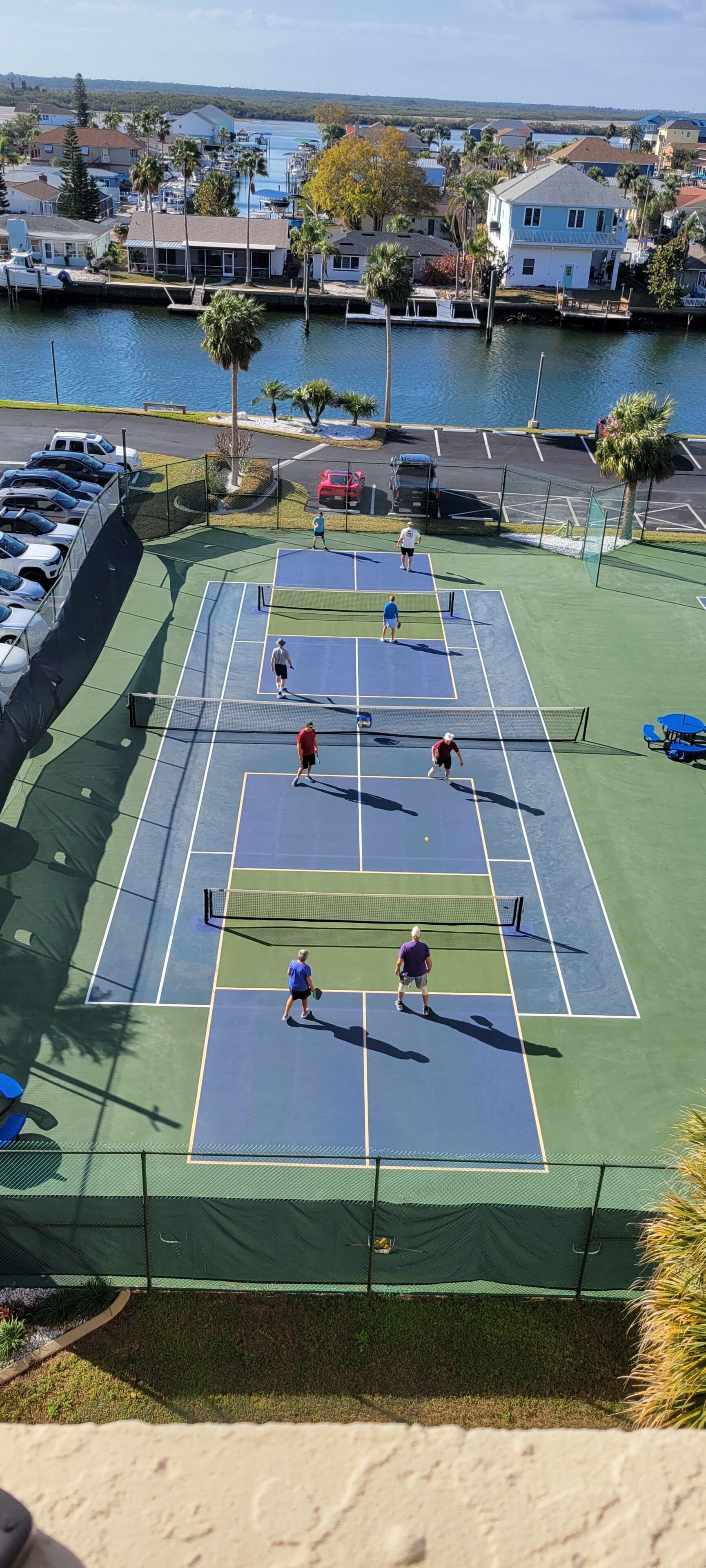 Una cancha de tenis con jugadores, rodeada por un estacionamiento y una masa de agua.