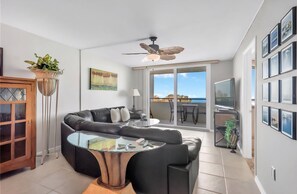 A modern living room with a sectional sofa, a glass coffee table, a ceiling fan, and a view of the outdoors through large windows.