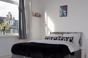 A bedroom with a bed, a window with curtains, a vase with flowers, and a painting on the wall.