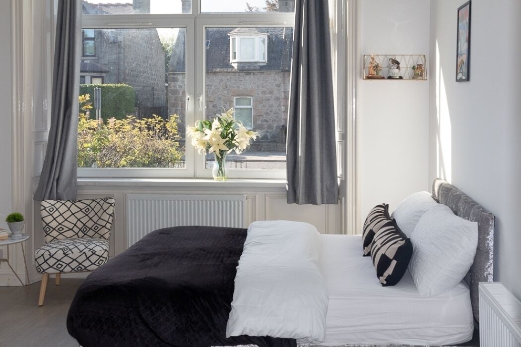 A bedroom with a bed, a chair, a window with curtains, and a vase with flowers.