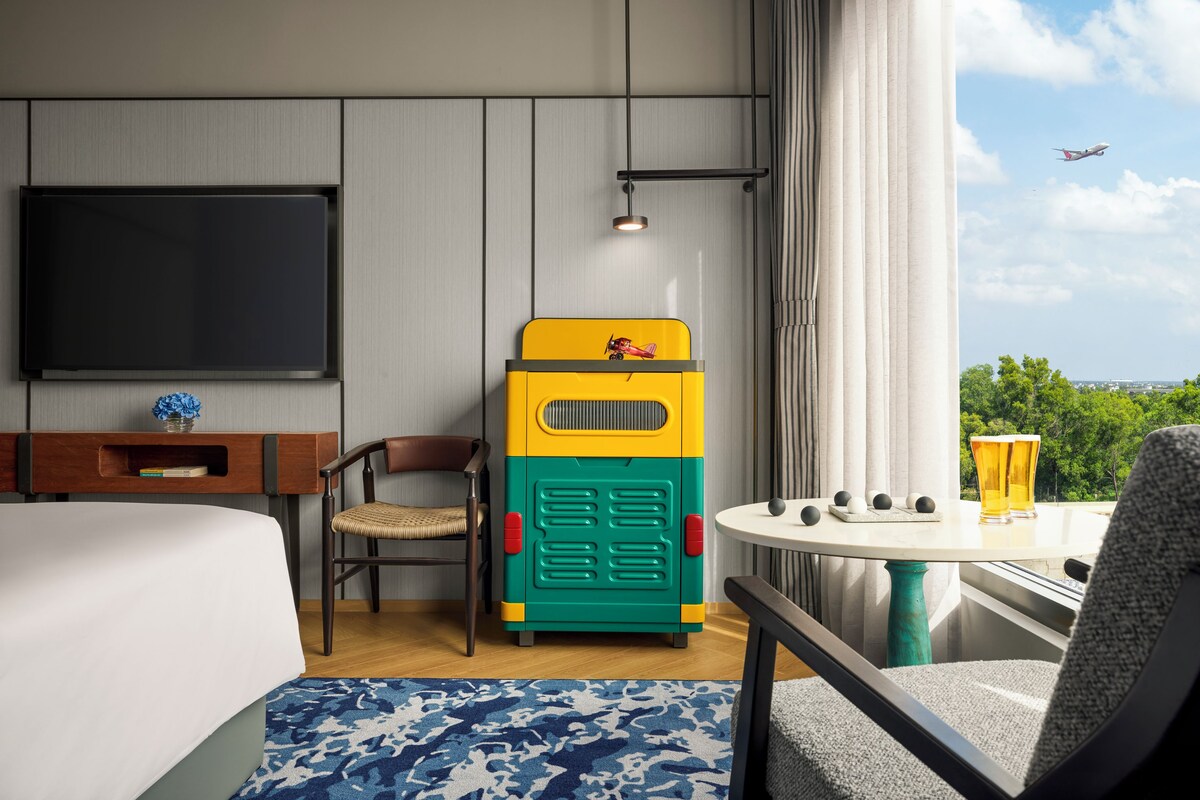 Habitación de hotel con una cama, un escritorio, una silla, un juguete de colores, una mesa con bebidas y vistas al cielo y al verdor.
