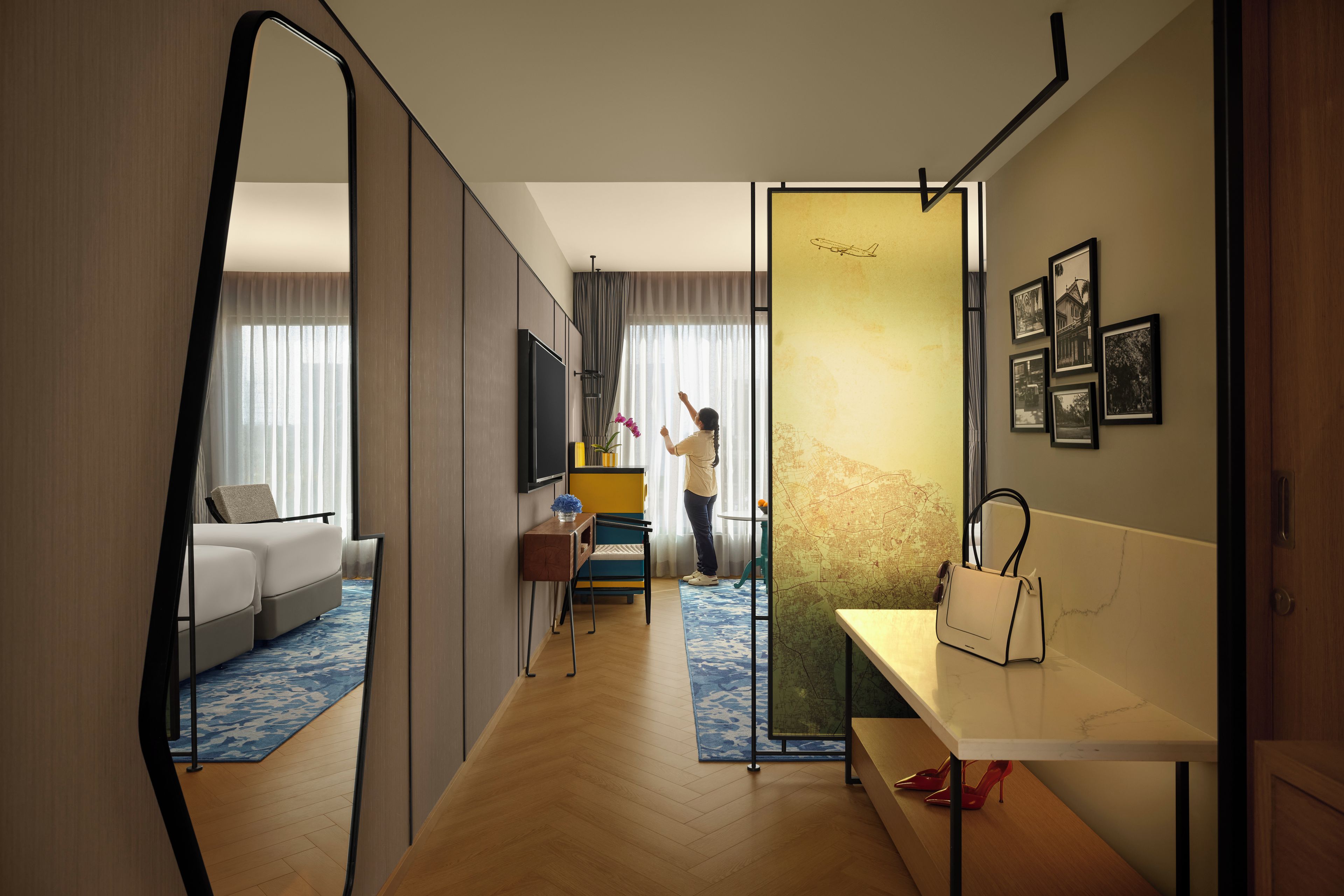 Une chambre d’hôtel avec un grand miroir, un canapé, une petite table avec un vase et un lit recouvert d’un tapis à motifs bleus et blancs.