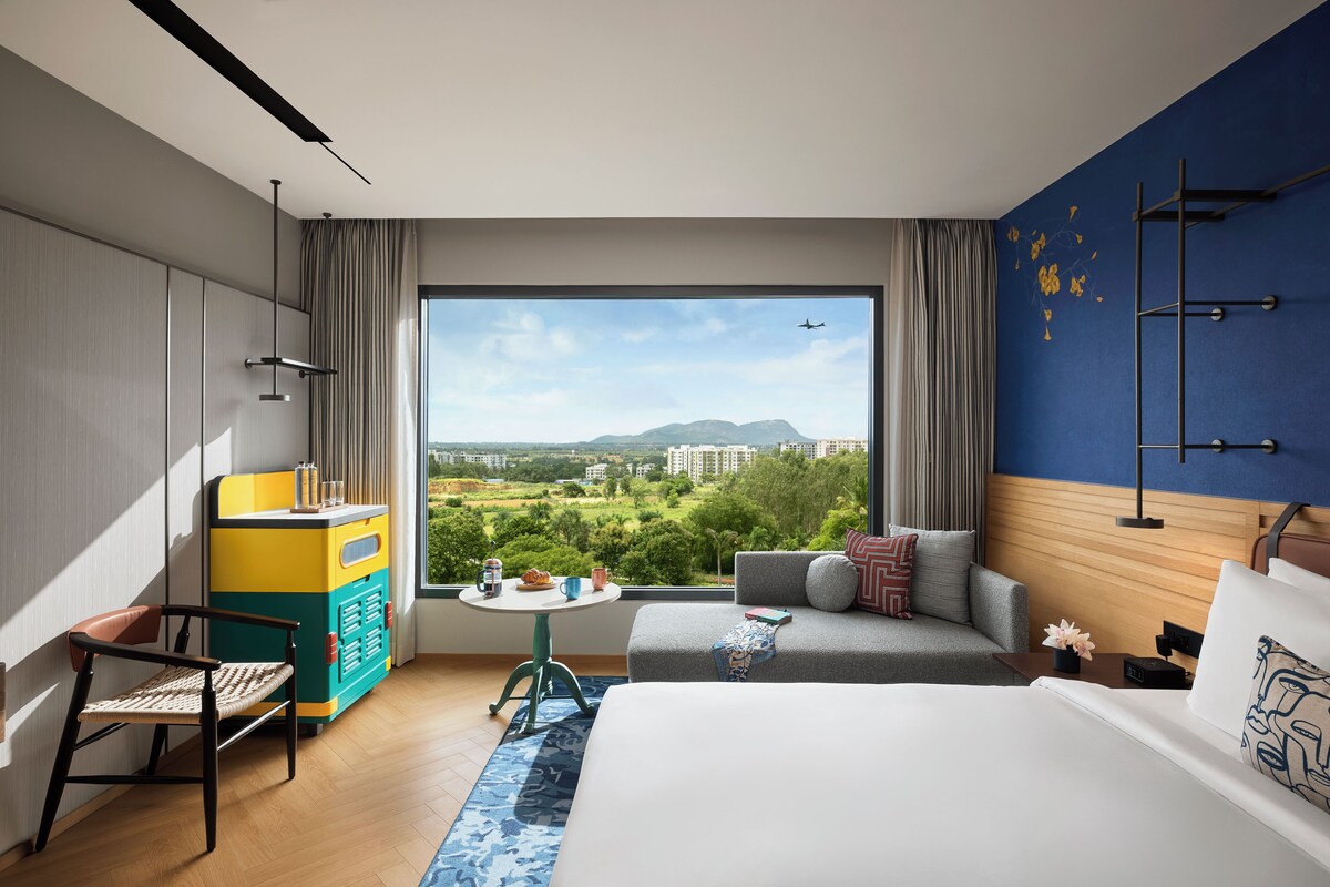Una habitación de hotel moderna con un ventanal grande, una cama, un sofá, una mesita pequeña y un armario de colores.