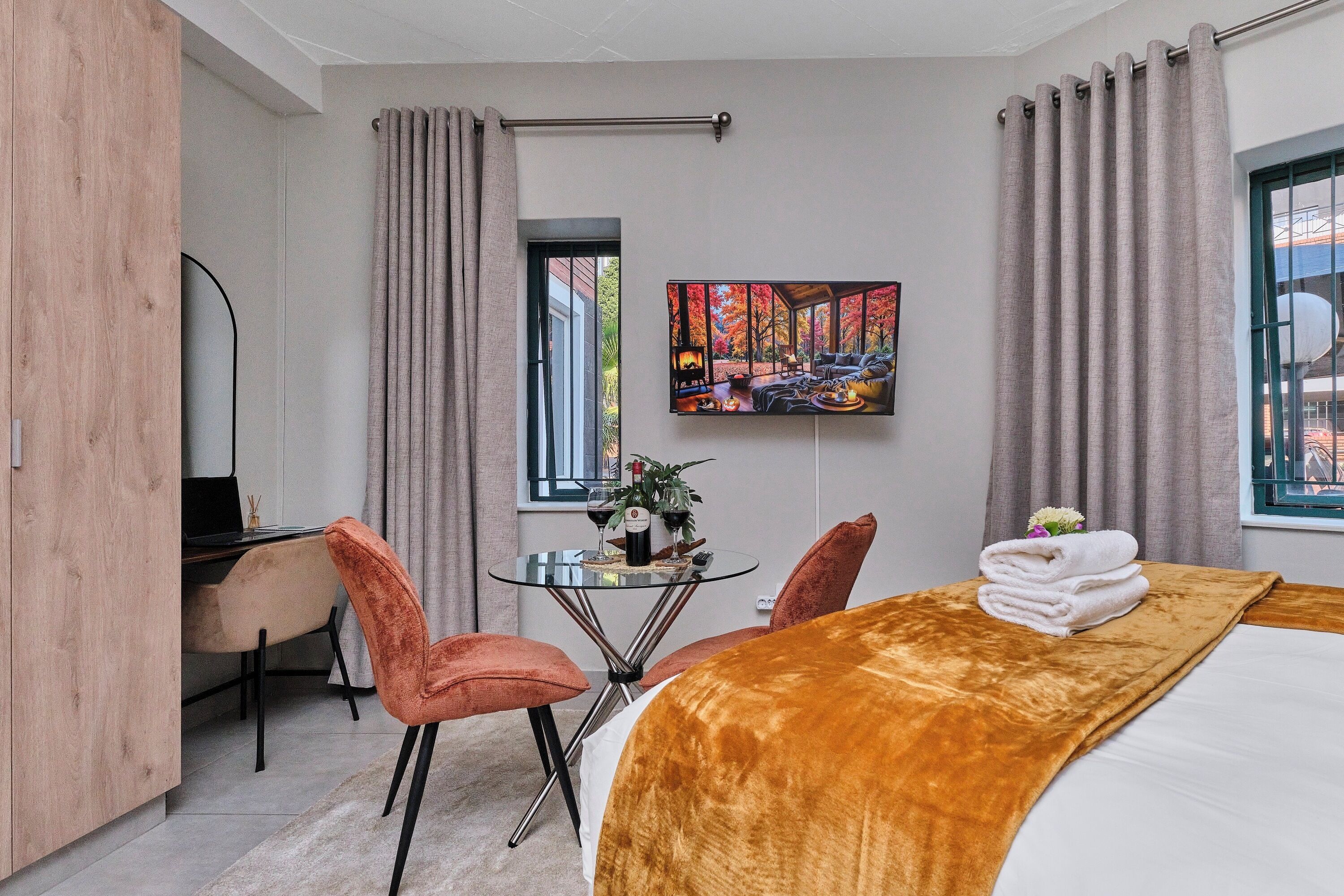 A hotel room with a bed, a desk, two chairs, a small table with a bottle of wine, and a painting on the wall.