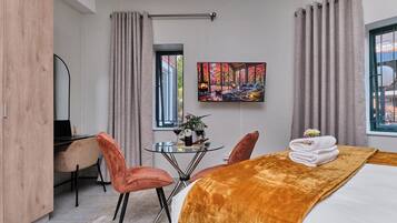 A hotel room with a bed, a desk, two chairs, a small table with a bottle of wine, and a painting on the wall.