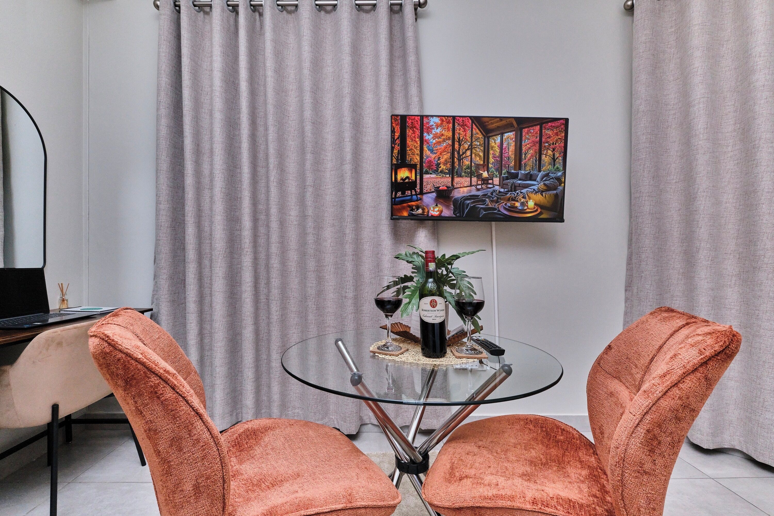 A modern living room with a glass table, two orange chairs, a painting on the wall, and a fireplace.