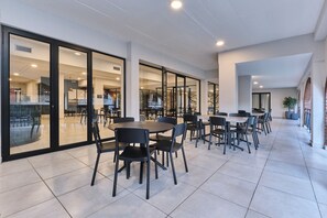 Amplio espacio interior con varias mesas y sillas, puertas de vidrio grandes que dan a una sala contigua, y piso de baldosas.
