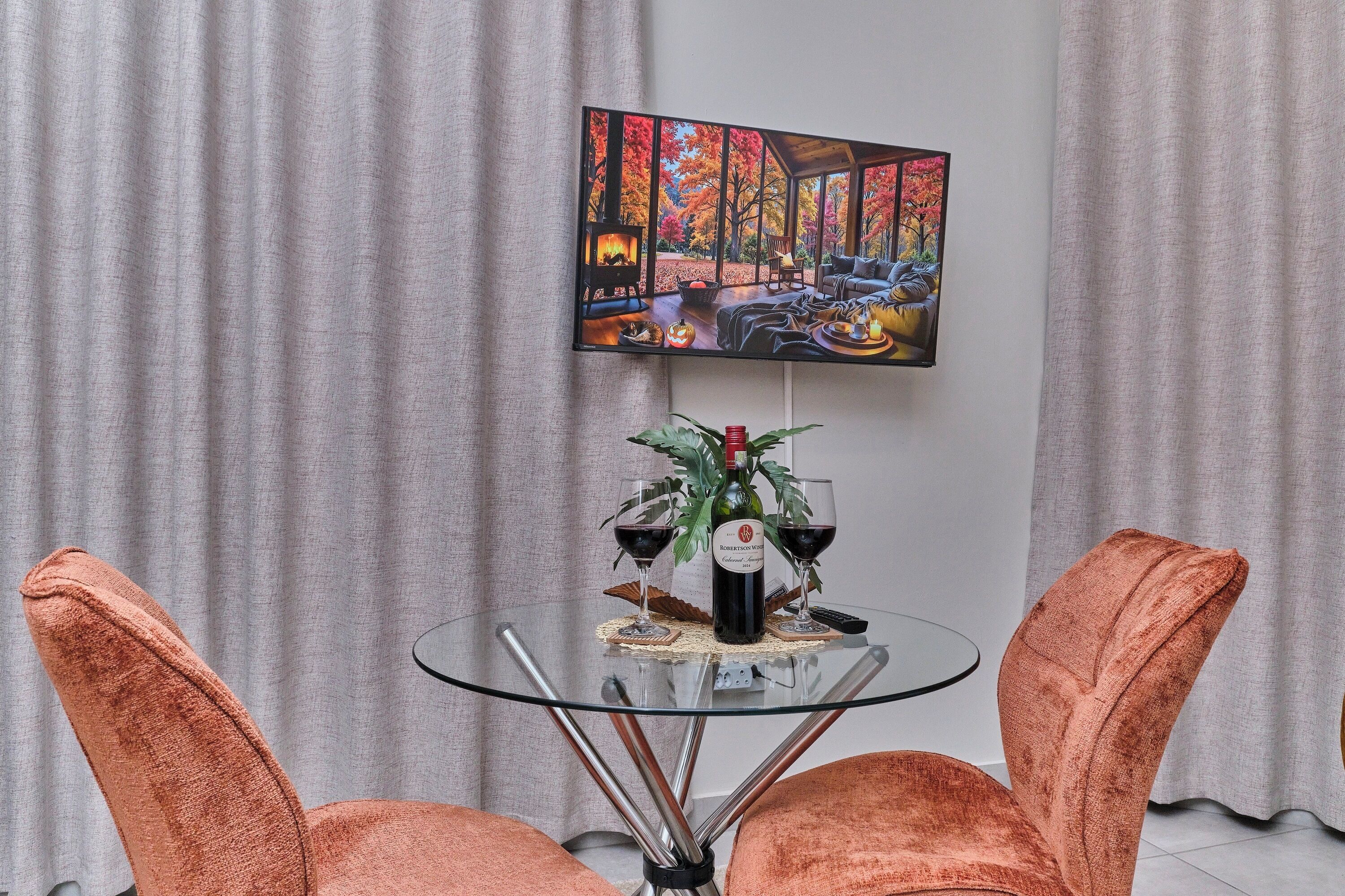 Un coin repas avec une table en verre, deux chaises orange, un tableau au mur et une bouteille de vin.