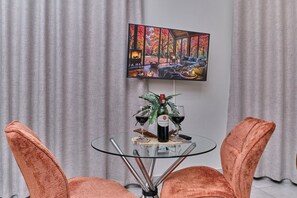 Een eethoek met een glazen tafel, twee oranje stoelen, een schilderij aan de muur en een fles wijn.