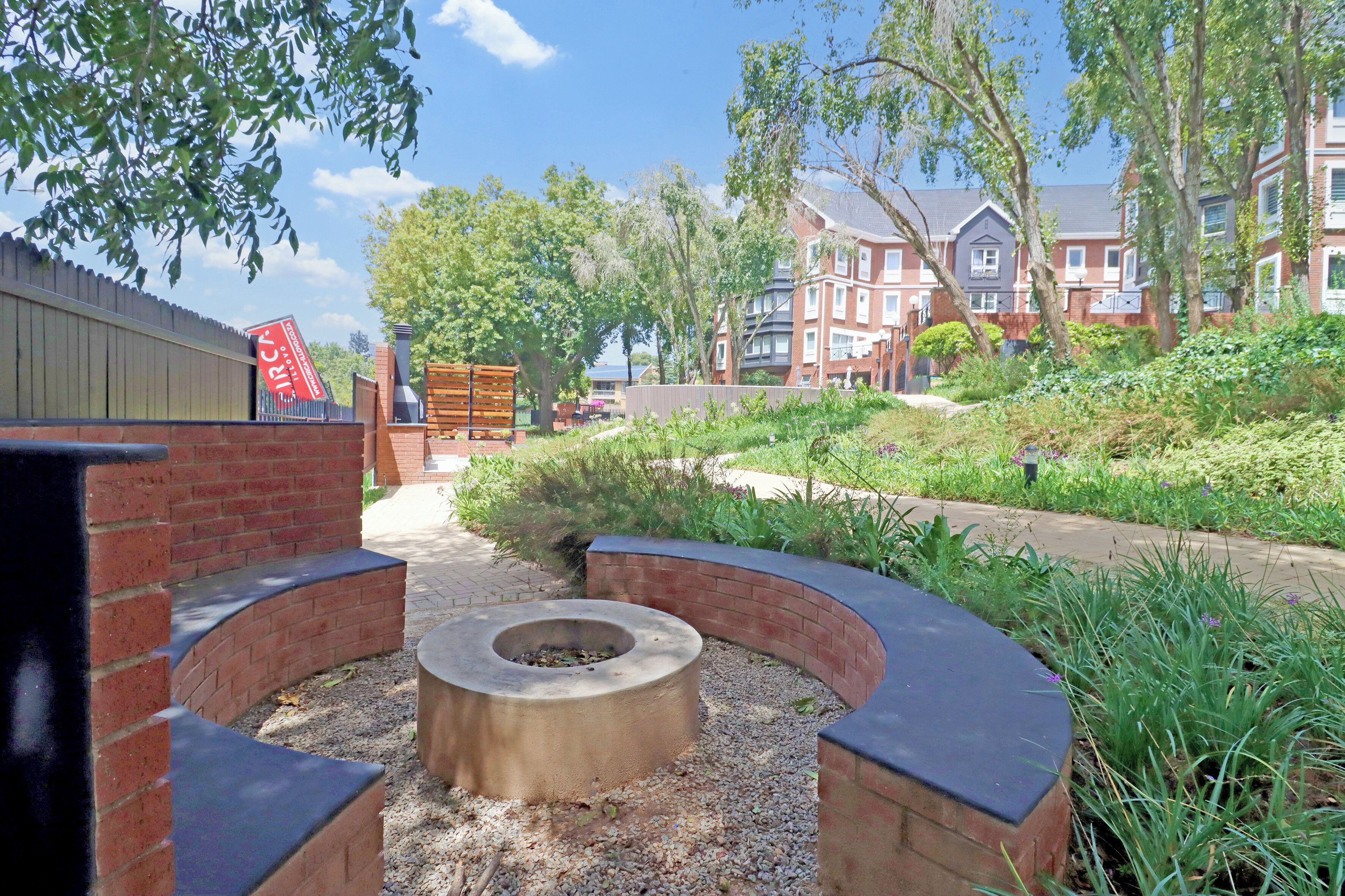 A courtyard with a circular seating area, a brick wall, and a pathway leading to a building with multiple windows.