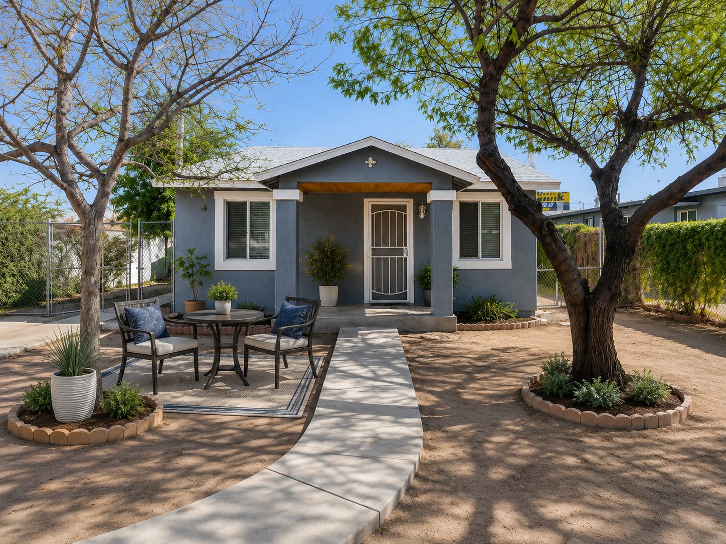 A single-story house with a front porch, a cross on the door, and a small garden with potted plants.
