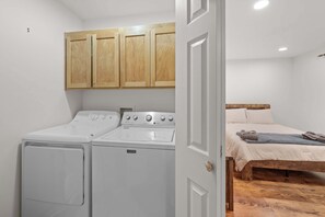 Una lavanderia con due lavatrici e un’asciugatrice, pensili in legno sopra, e un letto con testiera in legno nella stanza adiacente.