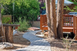 Un camí pavimentat que condueix a una terrassa de fusta amb una barbacoa, envoltada d'arbres i una tanca de fusta.