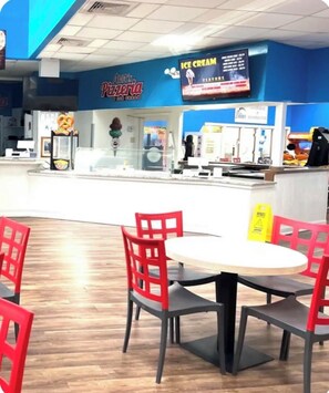 A food court with a pizza restaurant, a digital menu board, and red chairs around a table. - Oceanfront Coastal Comfort - Unique and Beautiful
Sand Dunes Resort (Myrtle Beach)