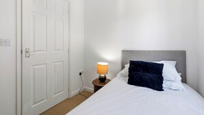 Ett hotellrum med en säng, ett nattduksbord med en lampa, en vit dörr och ett eluttag.