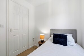 A hotel room with a bed, a nightstand with a lamp, a white door, and a power outlet. - Modern 3BR Retreat|Garden & Parking (Solihull)