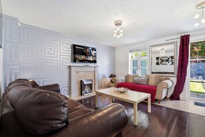 A living room with a fireplace, a television mounted on the wall, a sofa, a coffee table, and a window with curtains. - Modern 3BR Retreat|Garden & Parking (Solihull)