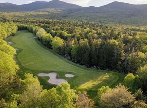 A golf course with a winding path, surrounded by dense forest and hills in the background.