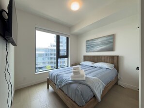 Ein Schlafzimmer mit einem Bett, einem Fenster, einem Gemälde und einem an der Wand montierten Fernseher.