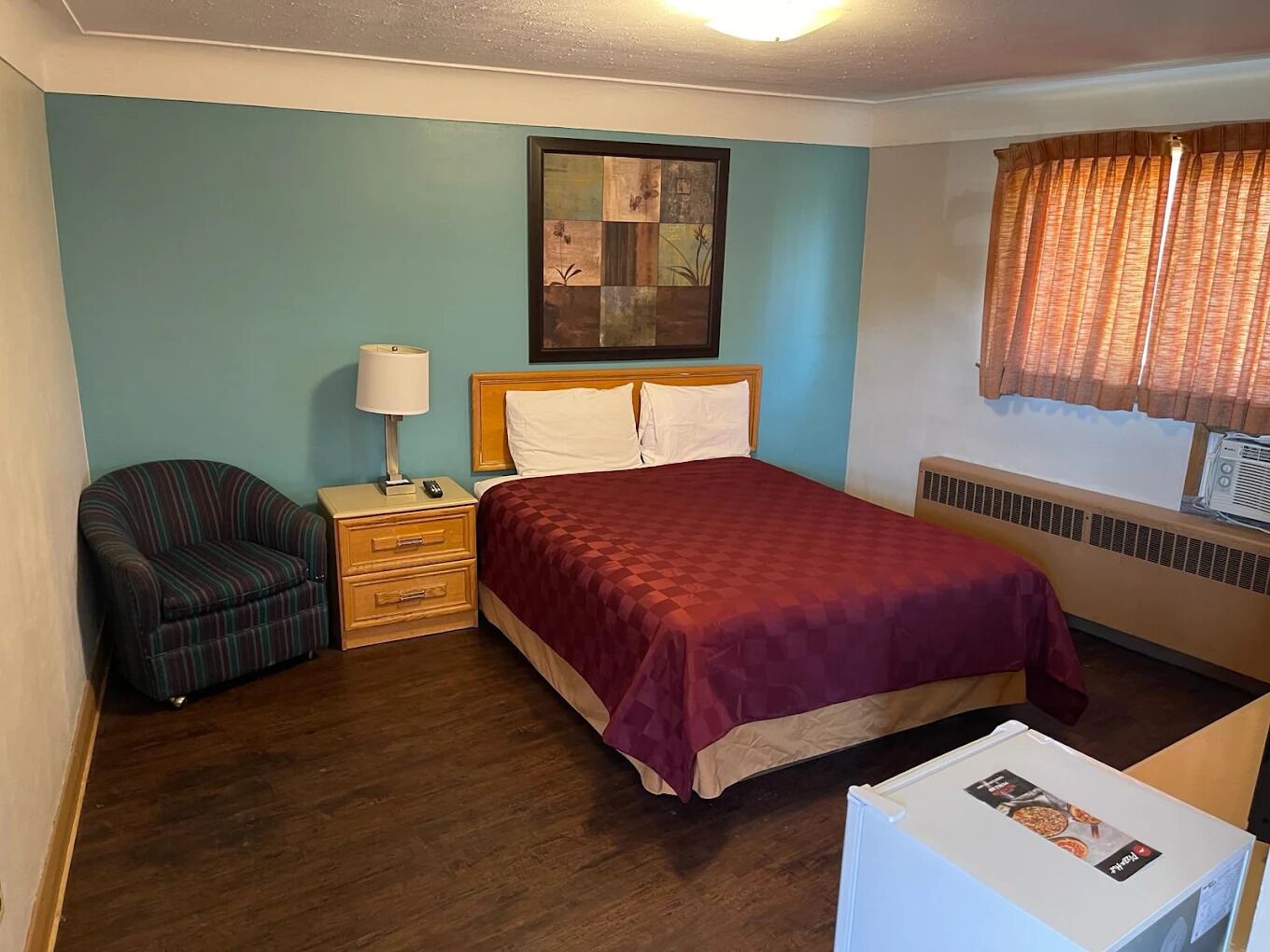 Una habitación de hotel con cama, una silla, una mesita de noche, una lámpara, un cuadro en la pared, una ventana con cortinas y un radiador.