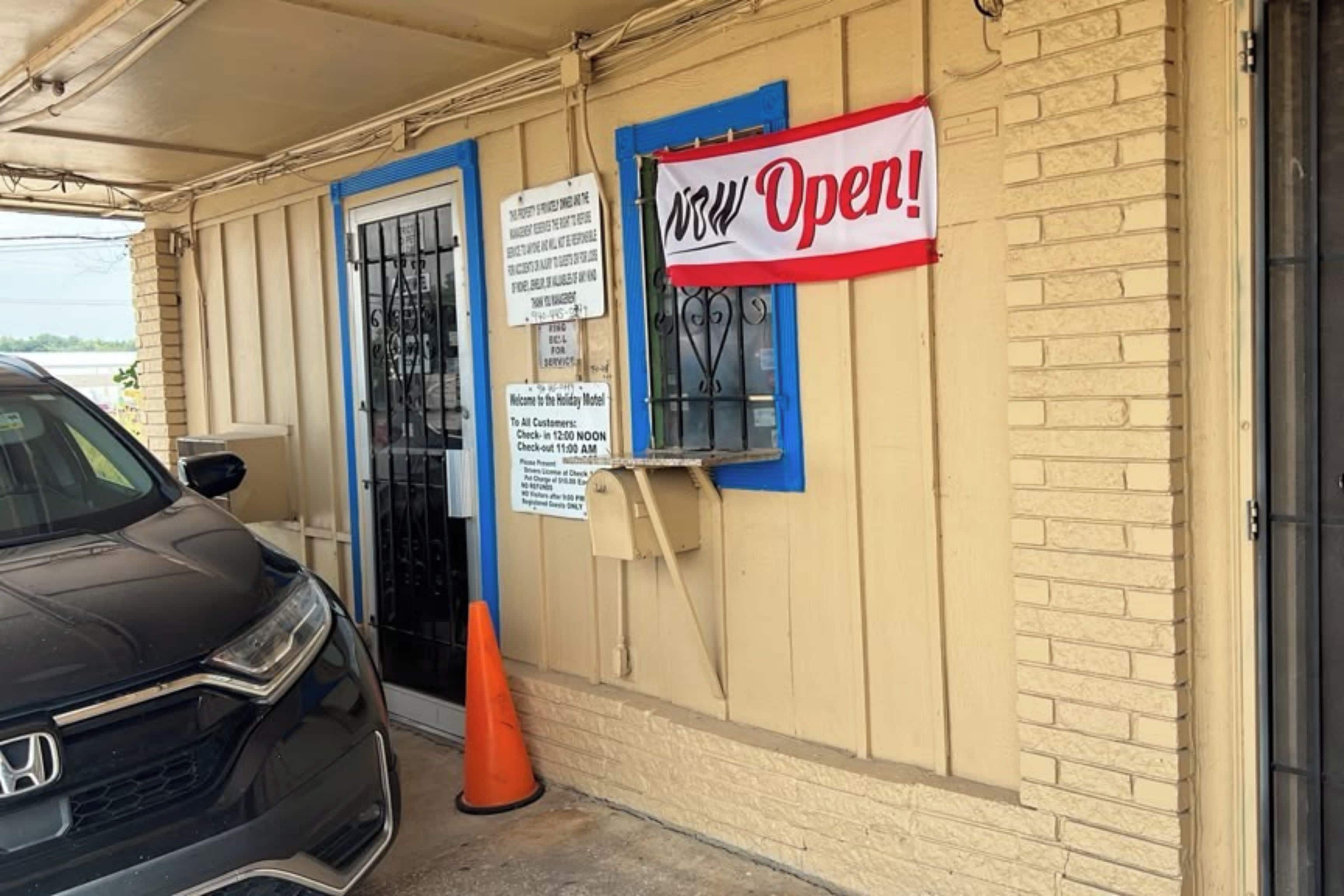 A hotel entrance with a 'View Open!' sign, a check-in time of 12:00 noon, and a parking area with a car and a traffic cone.