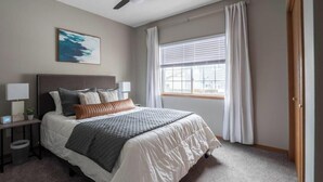 Ein Schlafzimmer mit Bett, Nachttisch, Lampe, einem Fenster mit Vorhängen und einem Deckenventilator.