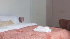 Ein ordentlich bezogenes Bett mit einer rosafarbenen Decke und zwei aufgerollten Handtüchern, ein brauner Stuhl und ein Nachttisch mit einer Lampe.