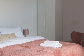A neatly made bed with a pink blanket and two rolled towels, a brown chair, and a bedside table with a lamp. - Studio Flat near University of Aberdeen (Aberdeen)
